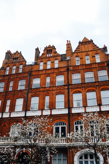 A multi-storey red brick building with white window frames and decorative architectural details, situated on a street with a pavement and two small trees in front. In the foreground, a moving trolley 