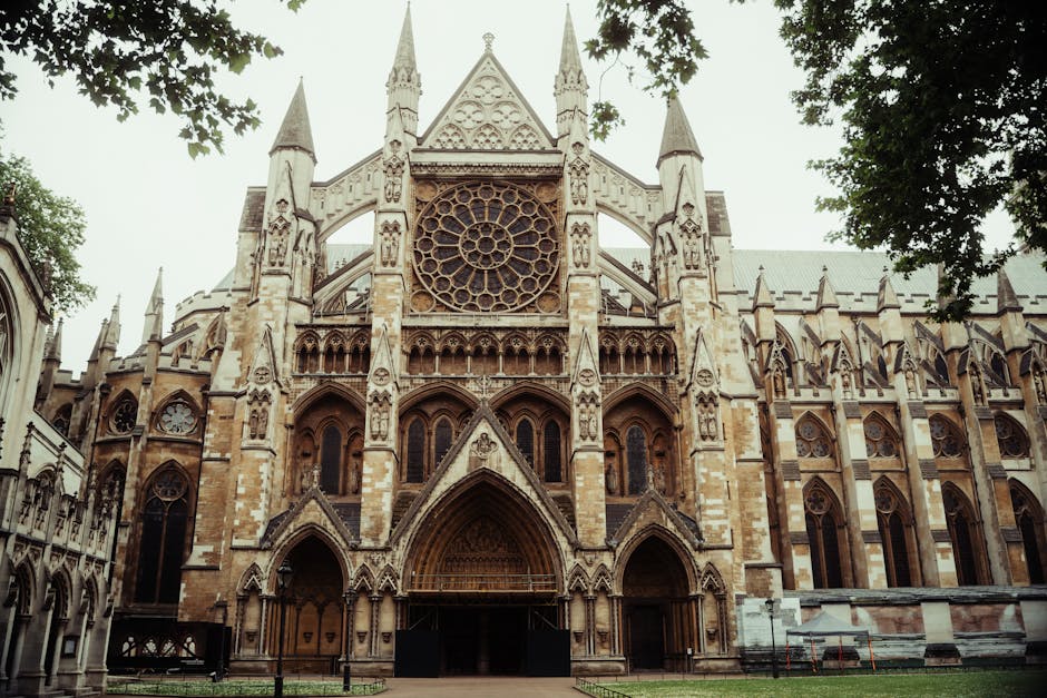 An exterior view of Westminster Abbey's historic Gothic façade with its two tall, ornate towers featuring decorative pinnacles and arched windows. The stonework displays intricate detailing, includin